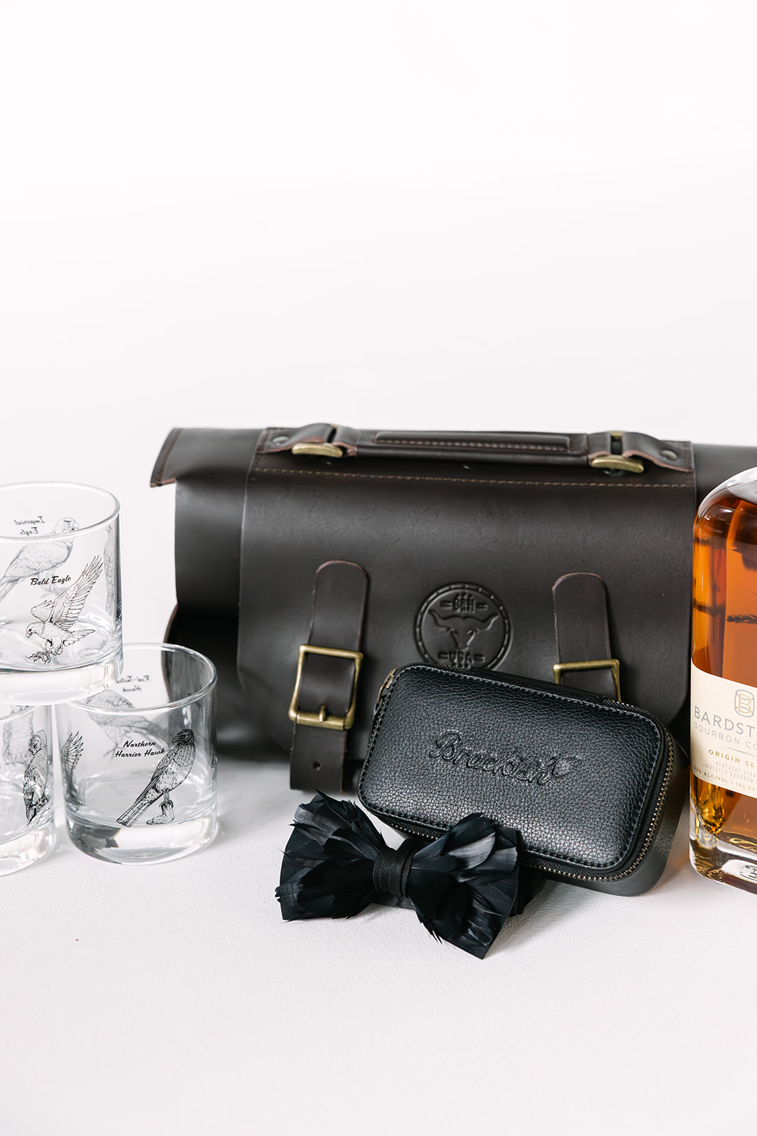 Dark leather bag with buckles, glass whiskey tumblers with bird illustrations, black leather zippered pouch, black feather bow tie, and a bottle of Bardstown bourbon.