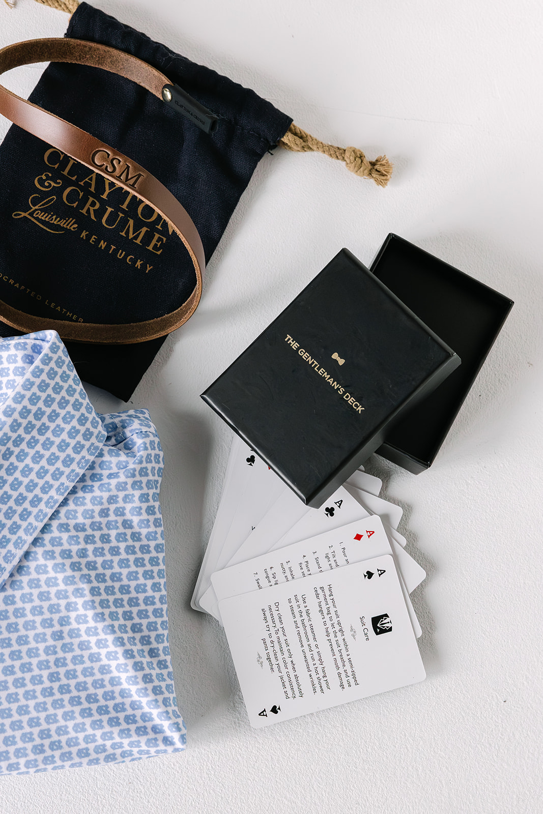 Black box labeled 'The Gentleman's Deck' with instruction cards spread out beside a blue patterned shirt and a leather bracelet on a black drawstring pouch.