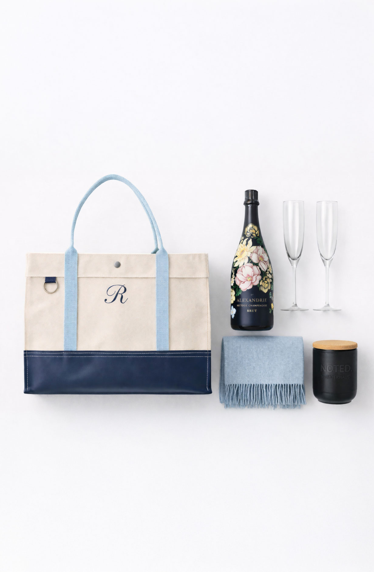 Flat lay of a beige and navy tote bag with a blue handle and letter R, a floral champagne bottle, two champagne flutes, a folded blue fringed scarf, and a black candle with a wooden lid.