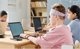Students focused on laptops in a bright classroom with shelves and a chalkboard in the background.