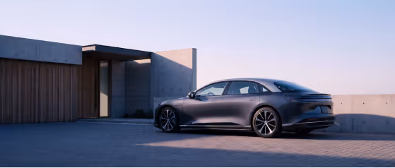 Sleek dark gray electric sedan parked in front of a modern concrete and wood house at sunset.