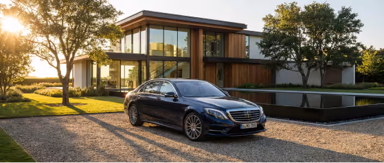 Black luxury sedan parked on gravel driveway in front of modern glass and wood house during sunset.