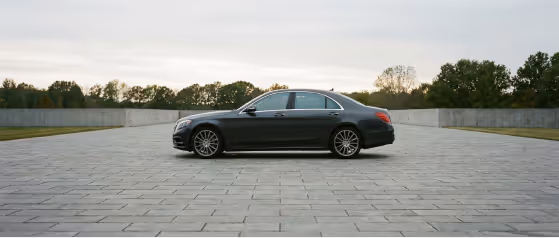 Black luxury sedan parked on a large paved stone area with trees and a cloudy sky in the background.