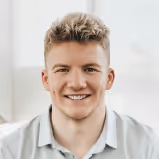 Young man with short blond hair smiling and wearing a light-colored shirt in soft indoor lighting.