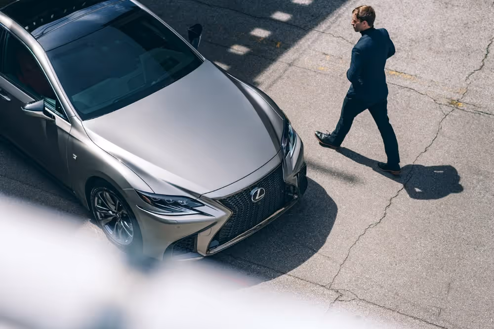 Man in dark clothing walking across cracked pavement beside a silver Lexus car parked on the street.