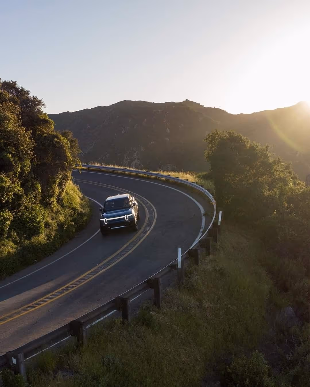 Electric SUV driving on a winding mountain road at sunset with hills and greenery surrounding.