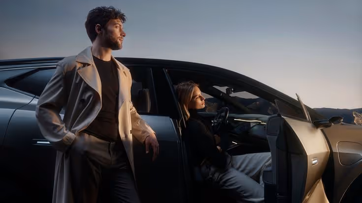 Man in beige trench coat standing beside an open car door with a woman sitting inside, both looking to the side during sunset.