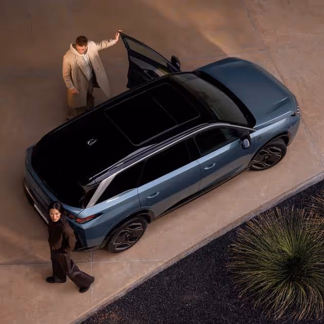 Overhead view of a blue SUV parked on a driveway with a man opening the driver's door and a woman standing nearby.