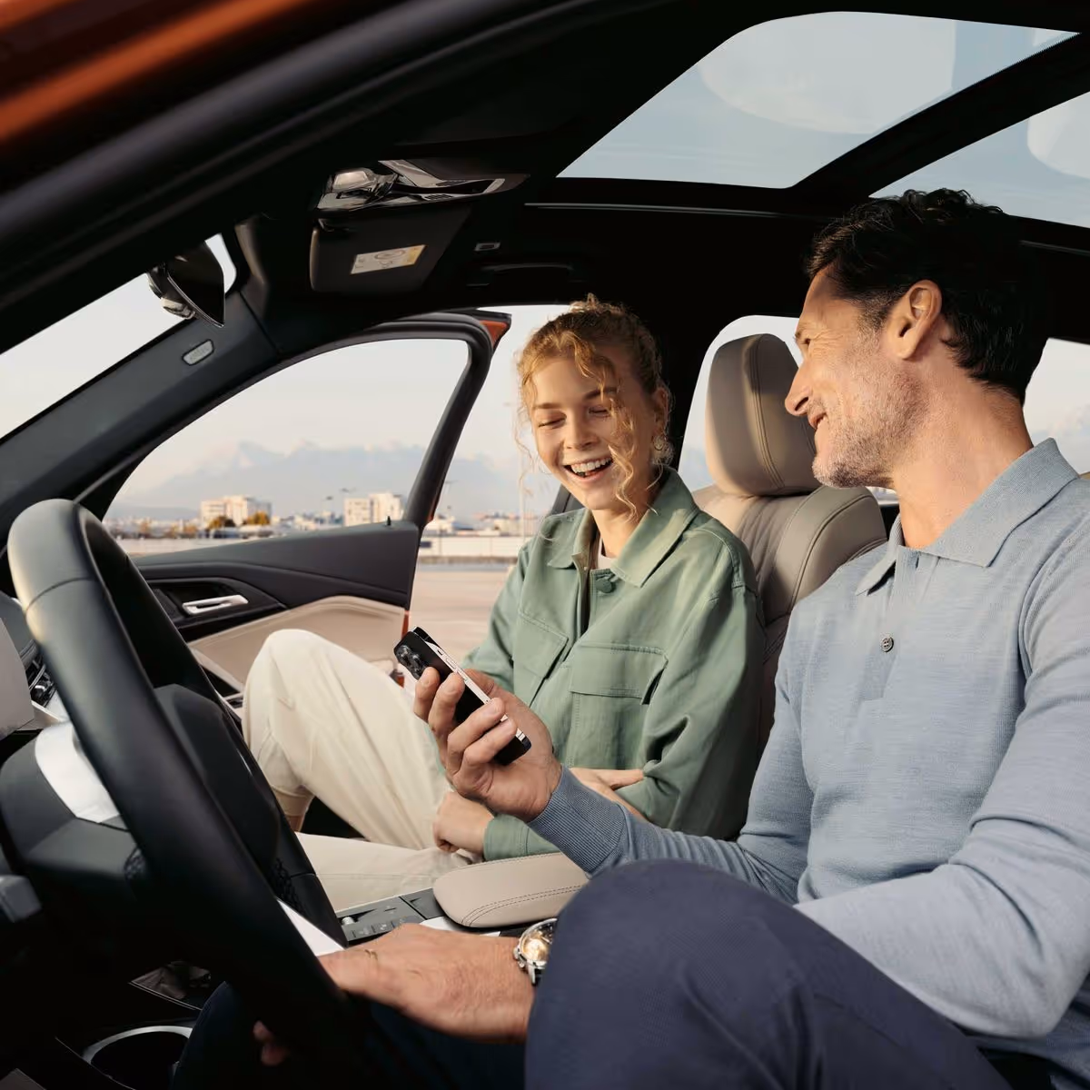Man and woman sitting in a car, smiling and looking at a smartphone together.