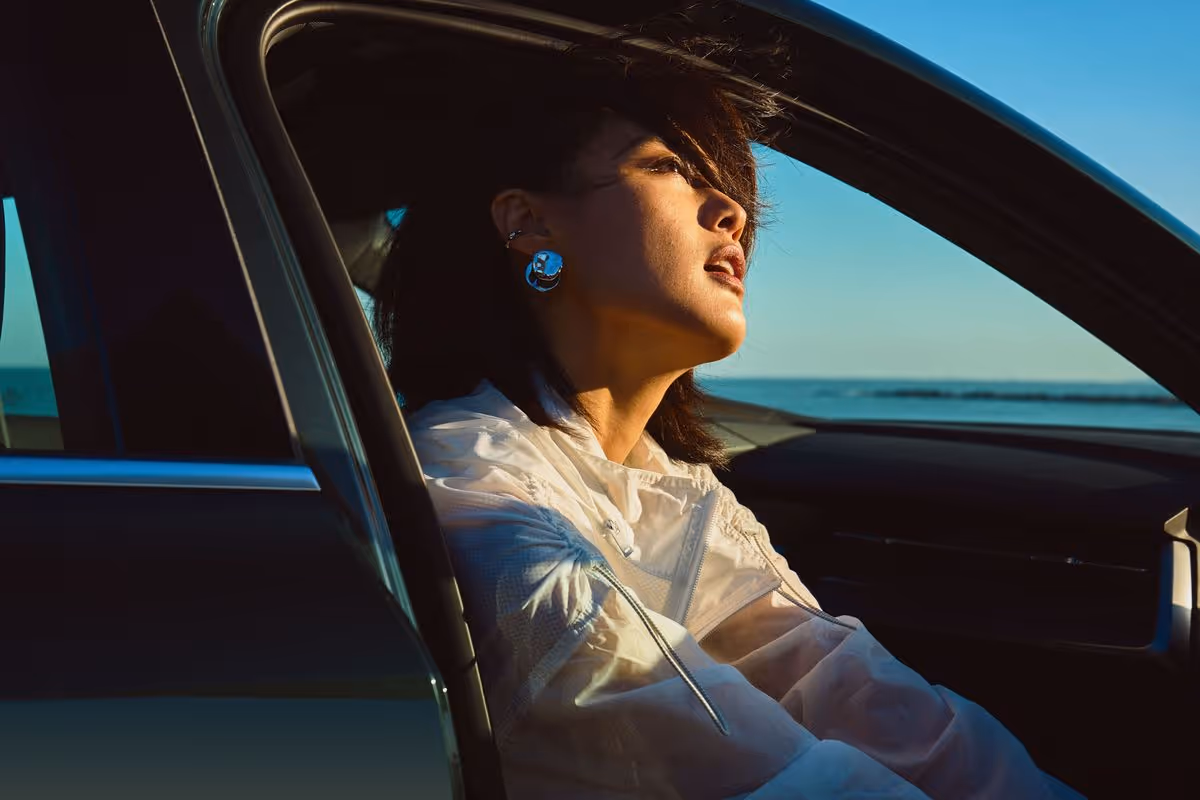 Woman with short dark hair and silver earrings looking out from a car window at sunset by the sea.