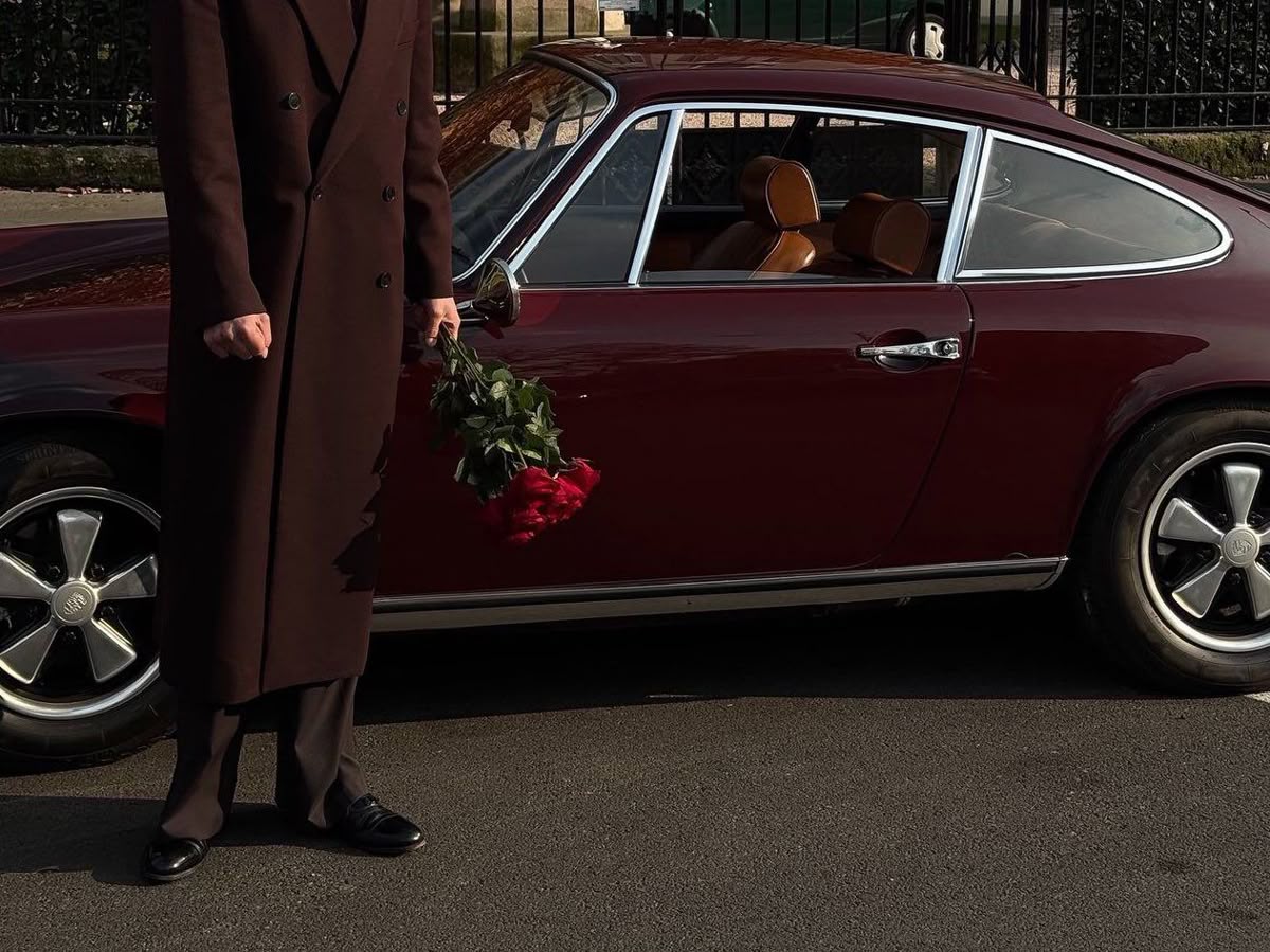 Person in a long dark coat holding a bouquet of red roses standing next to a vintage maroon car with tan leather interior.