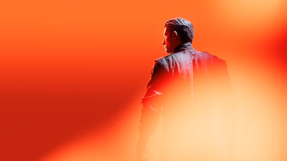 Man in a dark jacket looking to the side against an intense orange and red blurred background.