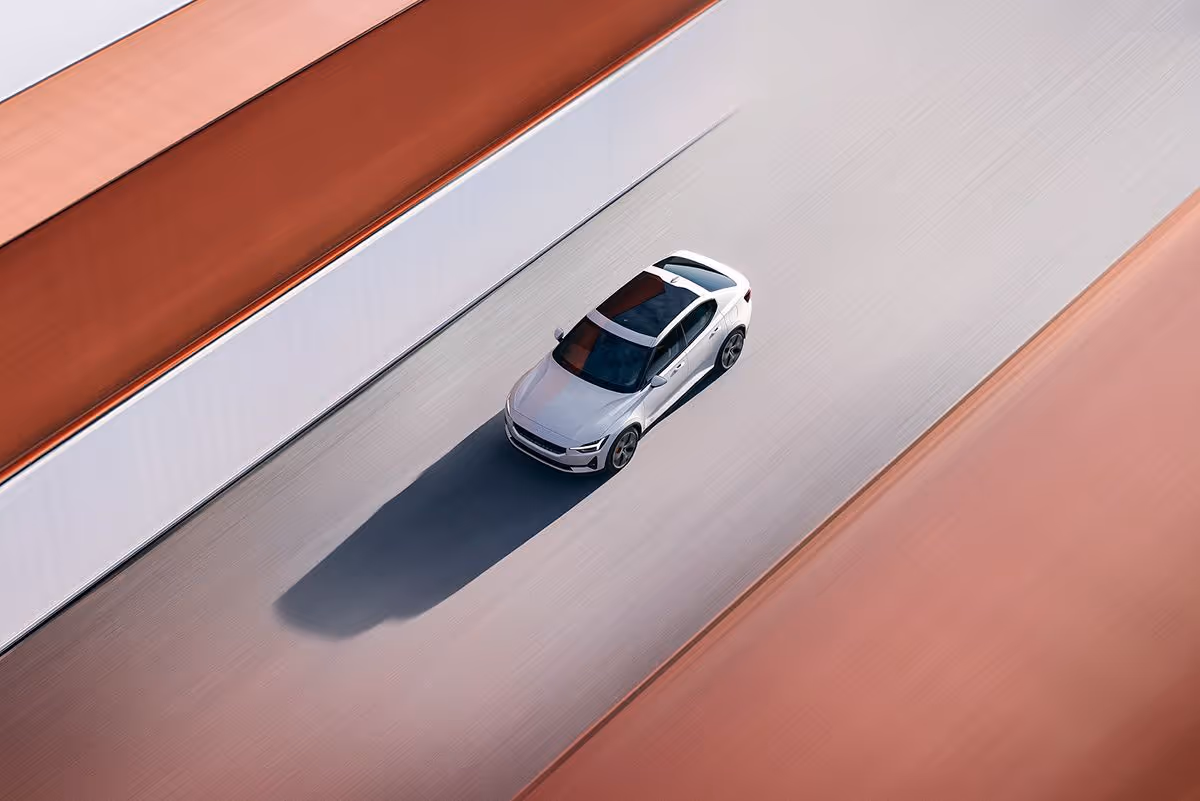 A silver car driving on a road bordered by orange and white walls, casting a long shadow.