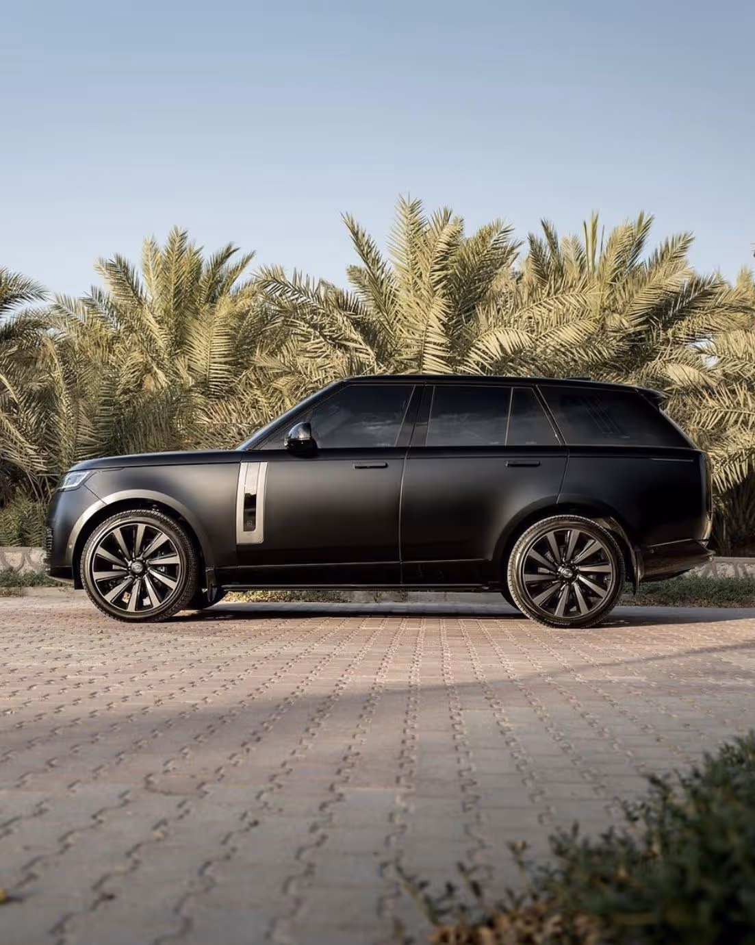 Black luxury SUV parked on patterned pavement with palm trees in the background under clear sky.