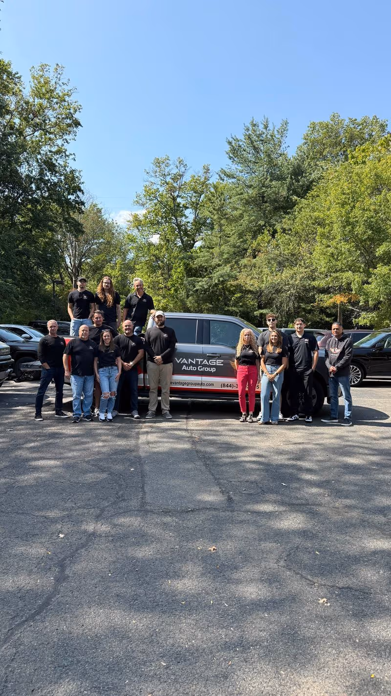 Group of 14 people standing in front of a Vantage Auto Group branded van in a parking lot surrounded by trees under a clear blue sky.