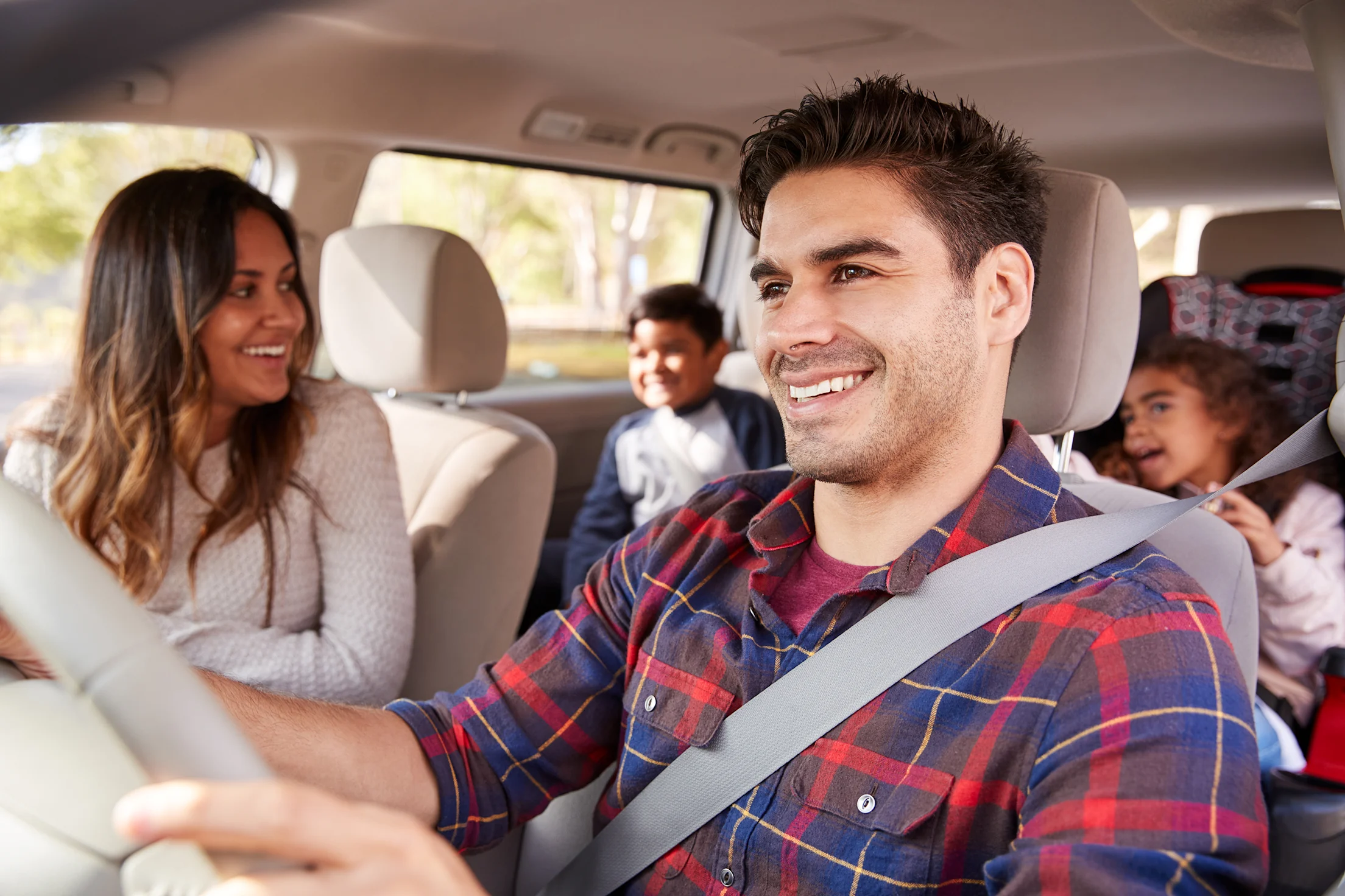 Smiling father driving a car with a mother in the front passenger seat and two children in the back seats.