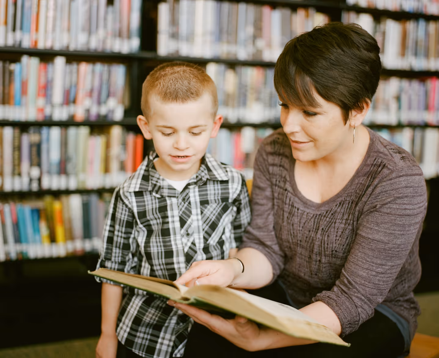 Woman reading a book with a young boy in a library with bookshelves in the background.