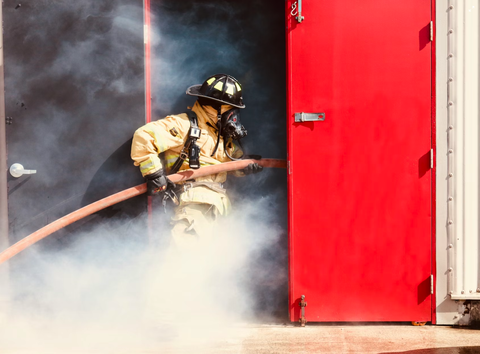 Firefighter in full gear holding a fire hose, standing in a doorway surrounded by smoke.