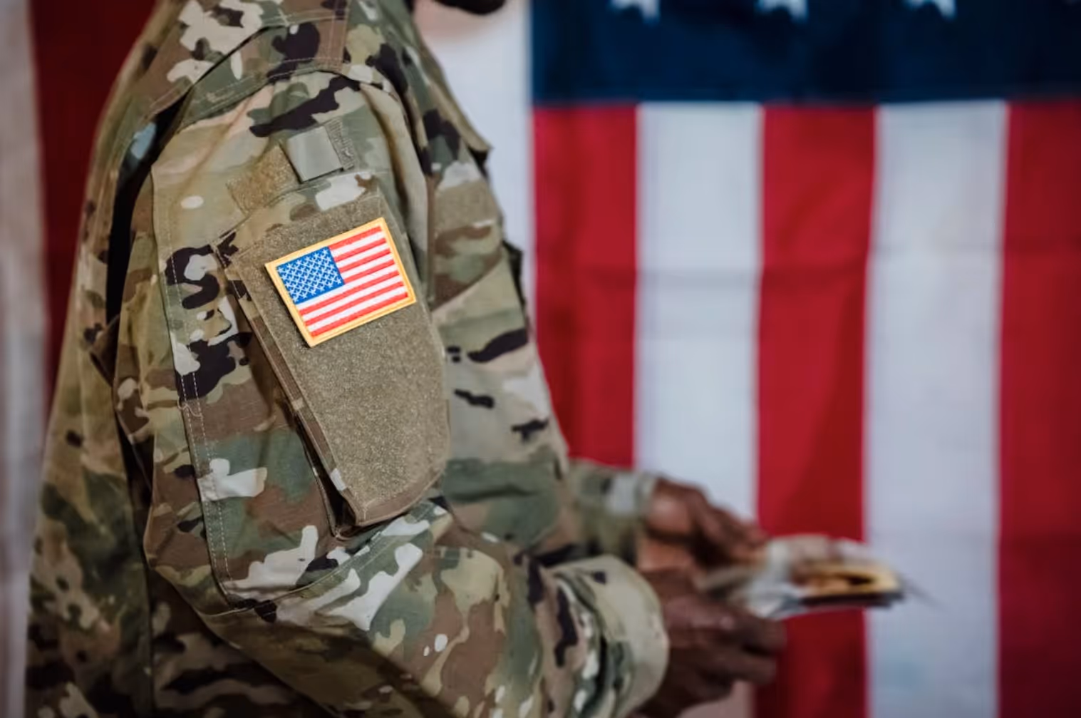 Close-up of a soldier in camouflage uniform with a US flag patch on the sleeve, holding papers with an American flag background.
