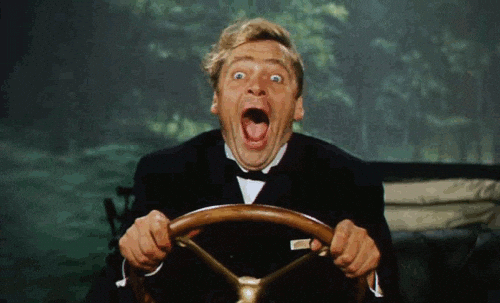 Man in suit gripping steering wheel with wide-eyed, open-mouthed frightened expression while driving.