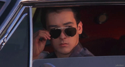 A man in a car lowering his sunglasses to look forward with a serious expression.