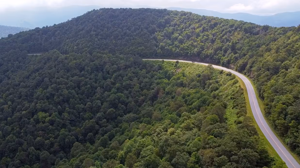 Winding road cutting through dense green forest on a mountain under a cloudy sky.