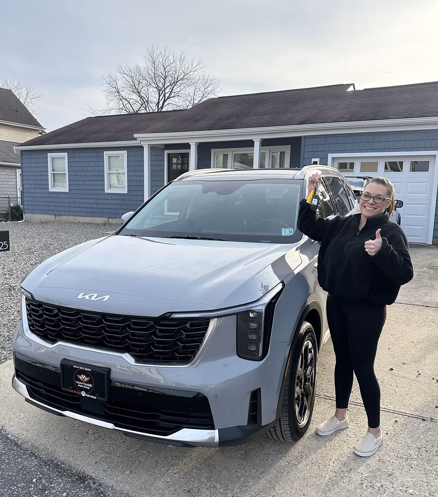 Smiling woman holding car keys and giving thumbs up standing next to a gray Kia SUV in front of a blue house.