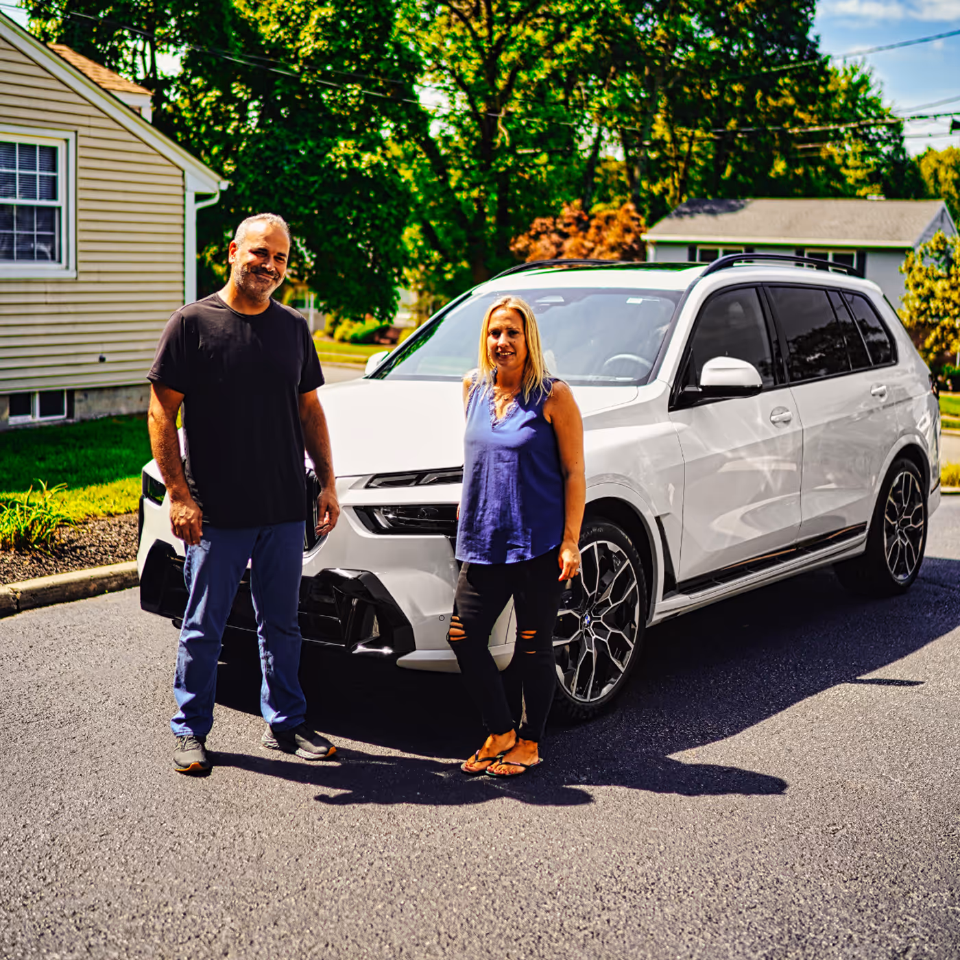 A man and a woman standing in front of a white SUV parked on a street in a sunny residential neighborhood.