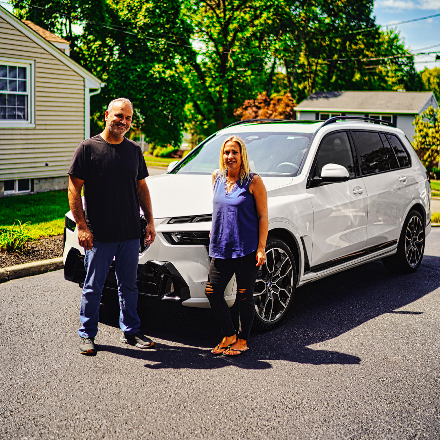 A man and a woman standing in front of a white SUV parked on a street in a sunny residential neighborhood.