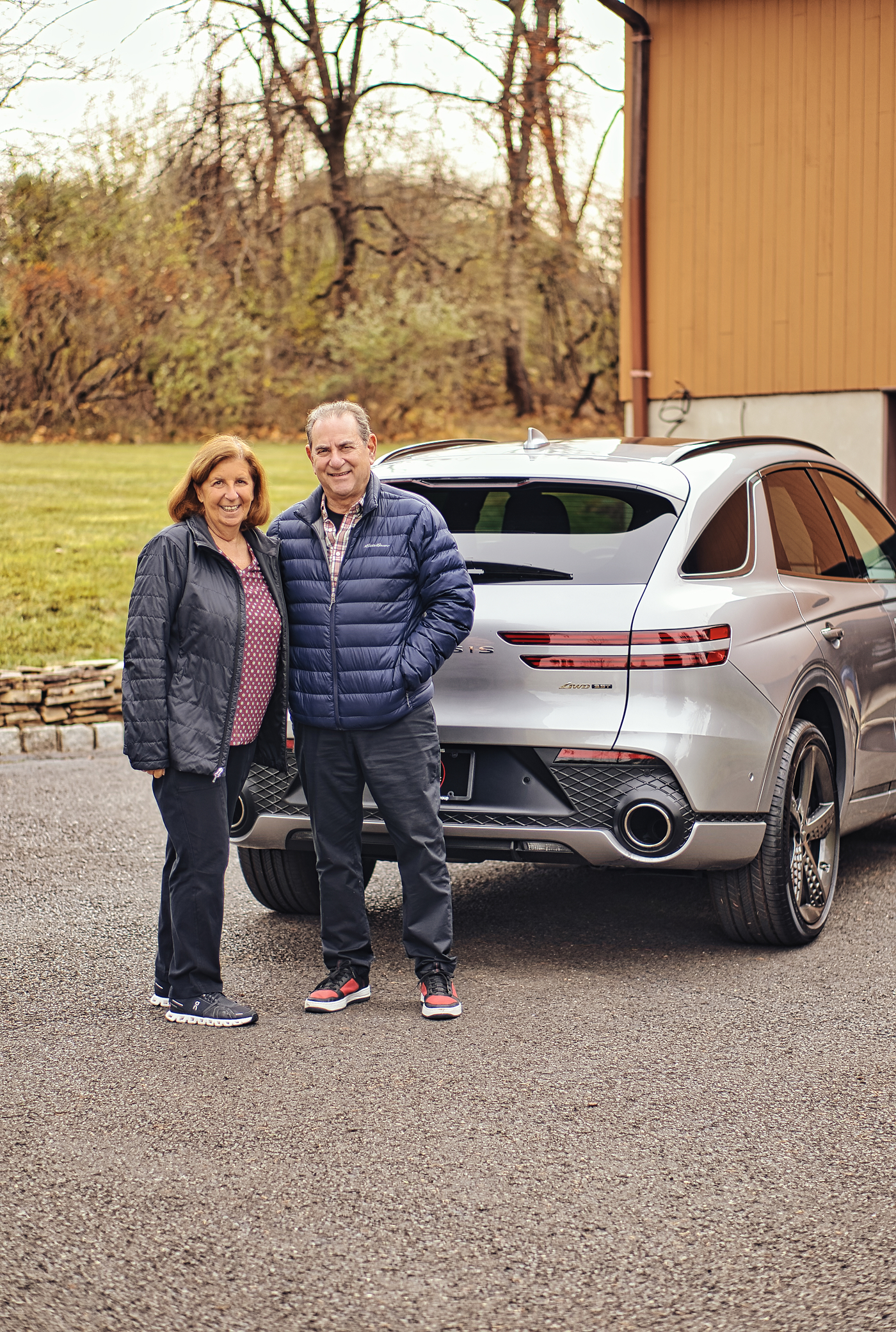Smiling middle-aged couple wearing jackets standing beside a silver luxury SUV in a driveway.