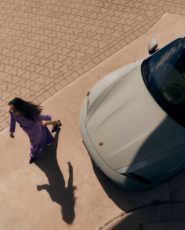 Top-down view of a woman in a purple dress walking beside a light-colored Porsche parked on a concrete and brick surface.