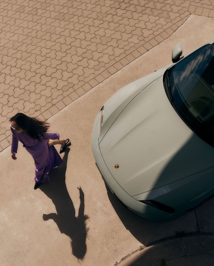 Top-down view of a woman in a purple dress walking beside a light-colored Porsche parked on a concrete and brick surface.