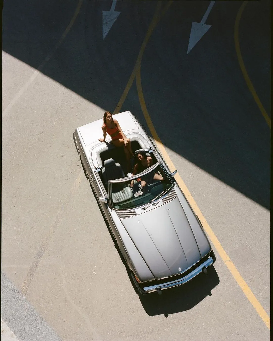 A silver convertible car on a road with a woman sitting on the back and another person driving.