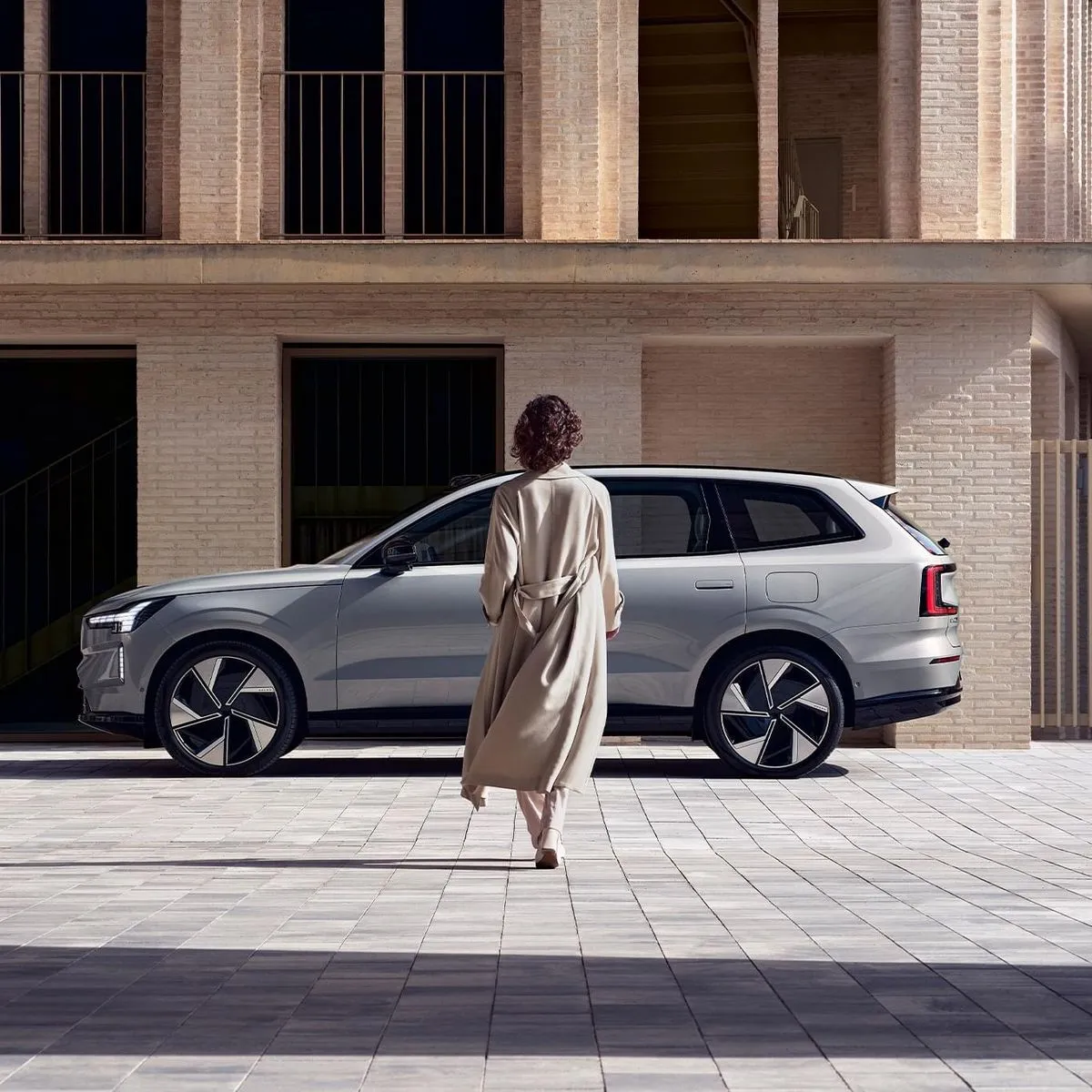 A person in a beige coat walking towards a silver modern SUV parked in front of a brick building.