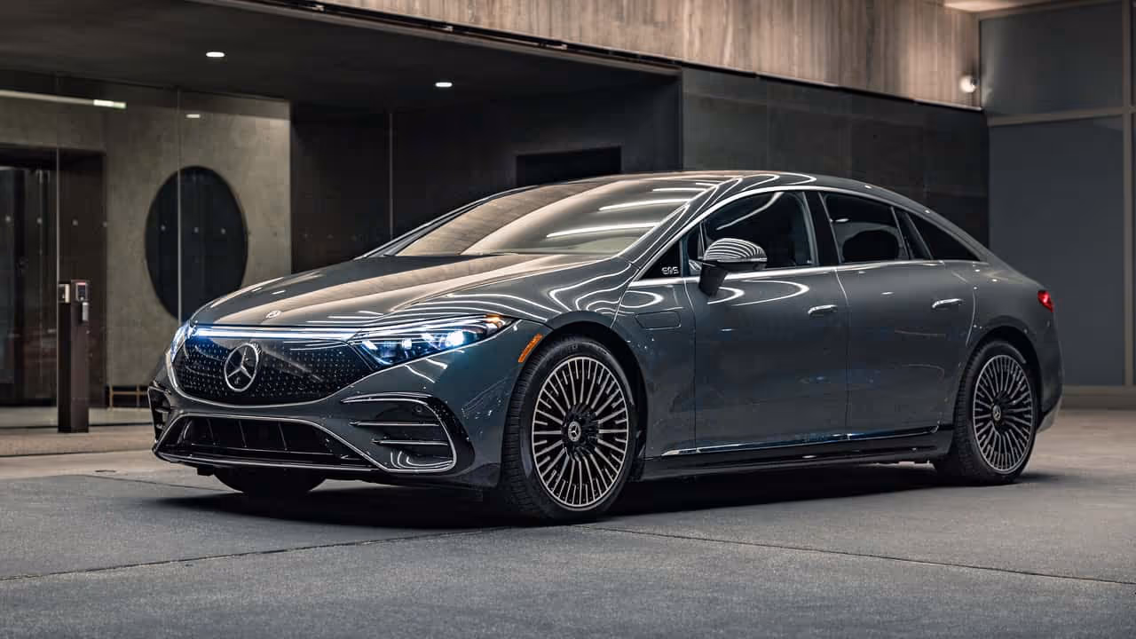 Sleek dark gray Mercedes-Benz EQS electric sedan parked in an indoor modern setting with headlights on.