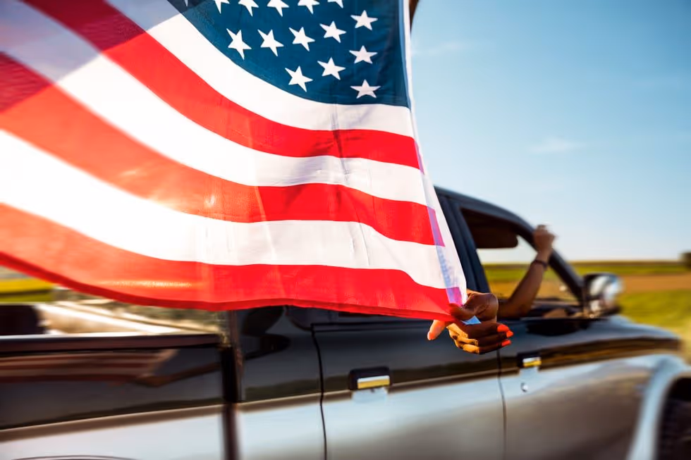 Person with orange nail polish holding an American flag out of a black pickup truck window.