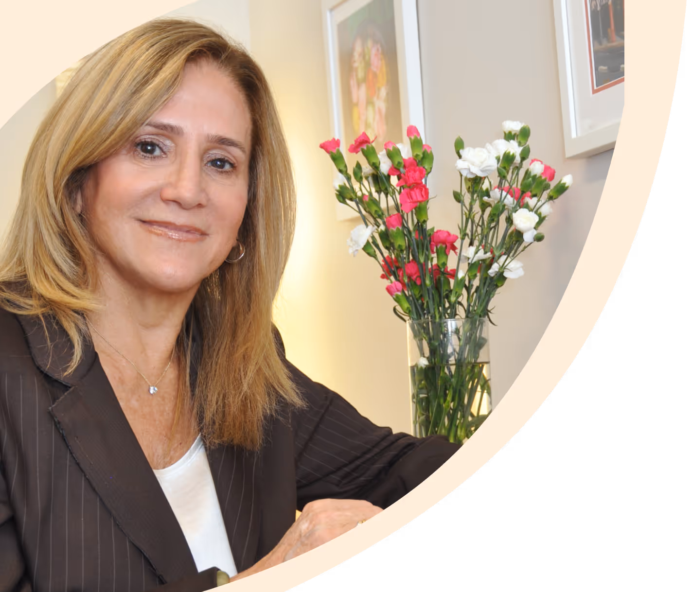 Woman with light brown hair wearing a dark blazer and white top with flowers in a vase behind her.