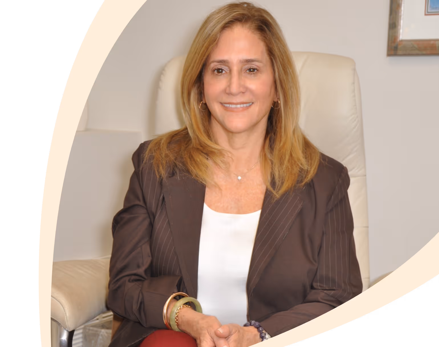 Woman with blonde hair in pinstripe blazer and white top sitting in a cream chair smiling.
