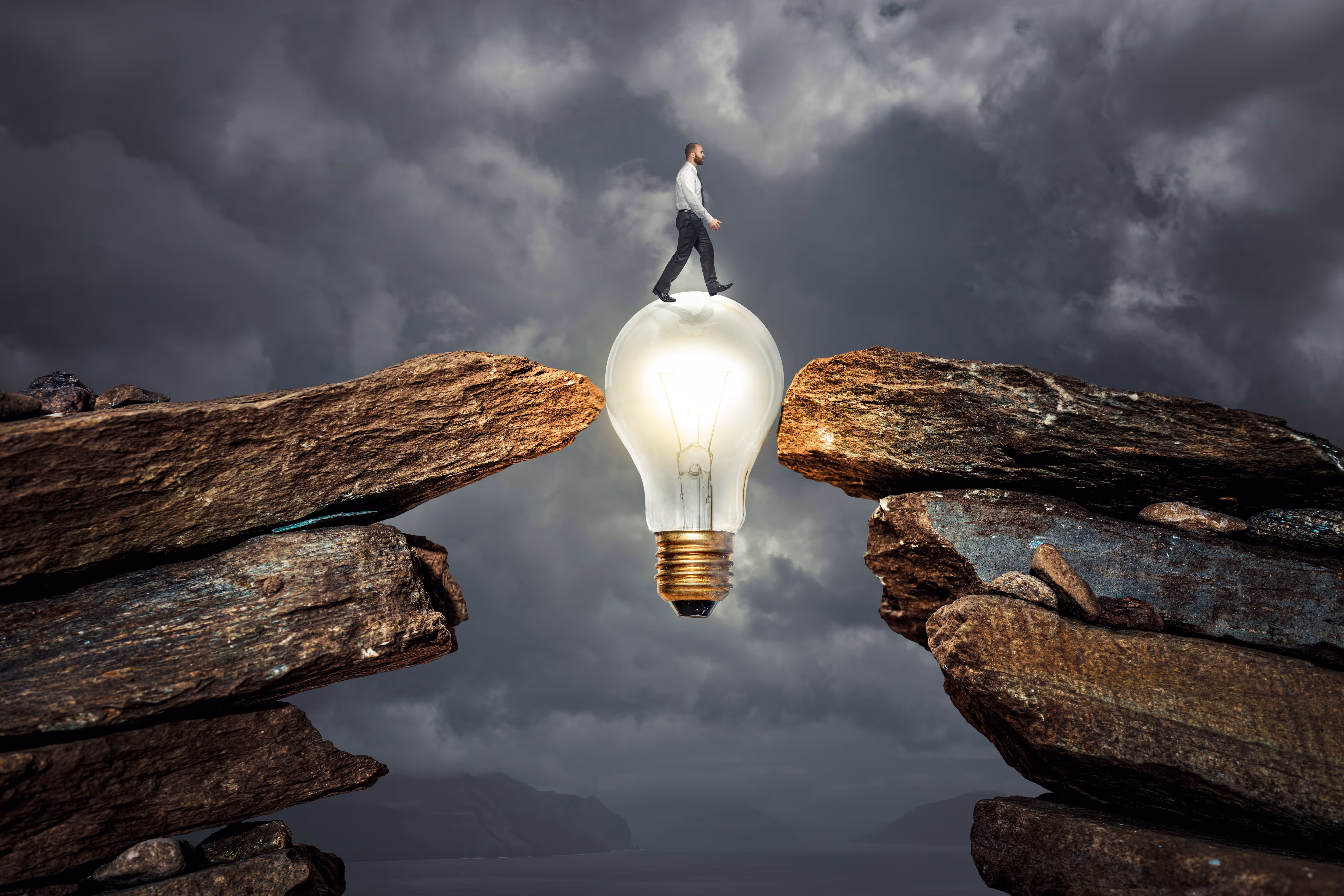 Man in business clothes walking on a glowing light bulb bridging two rocky cliffs under a cloudy sky.