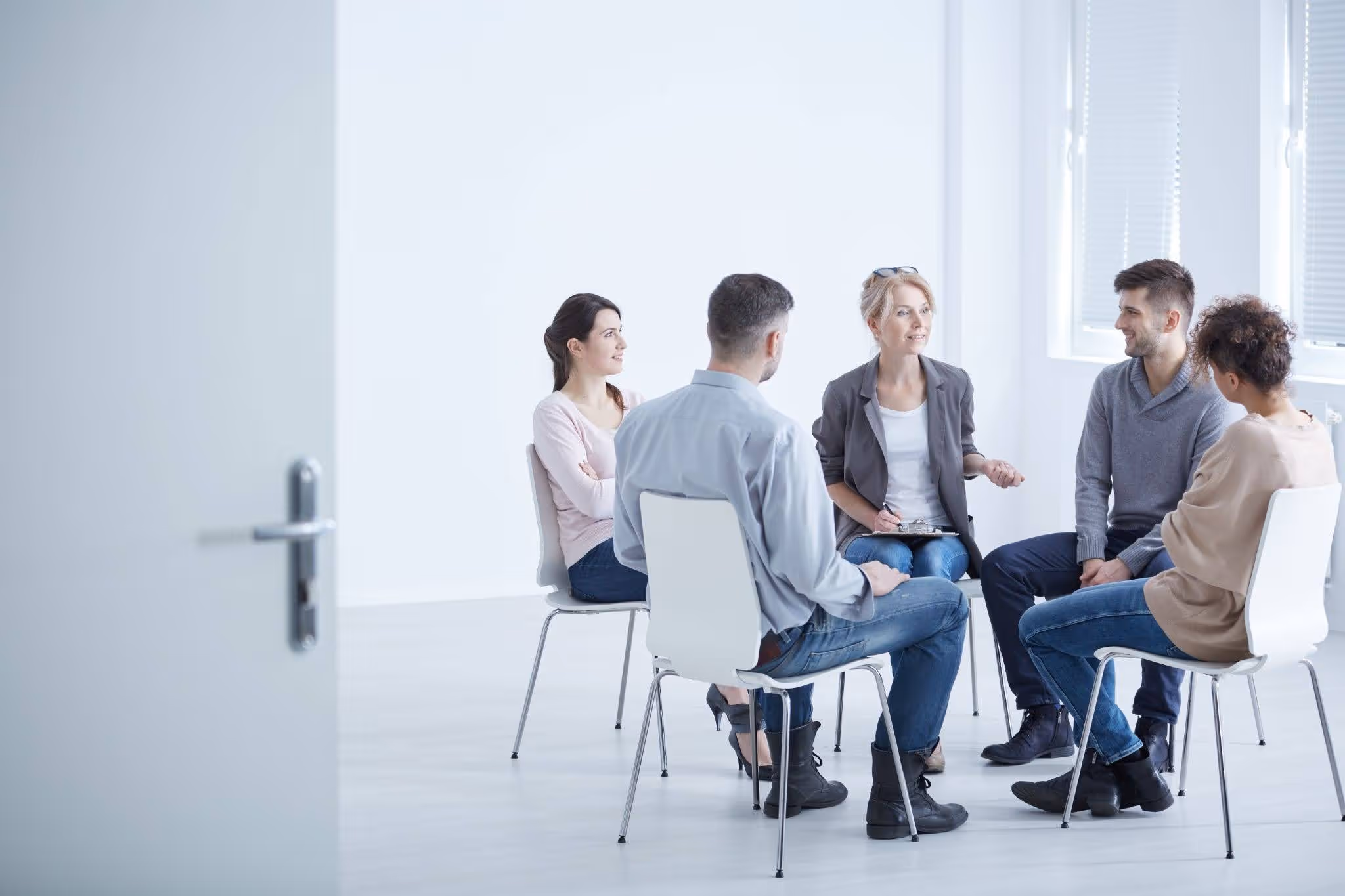 Five people seated in a circle in a bright room having a group discussion or therapy session.