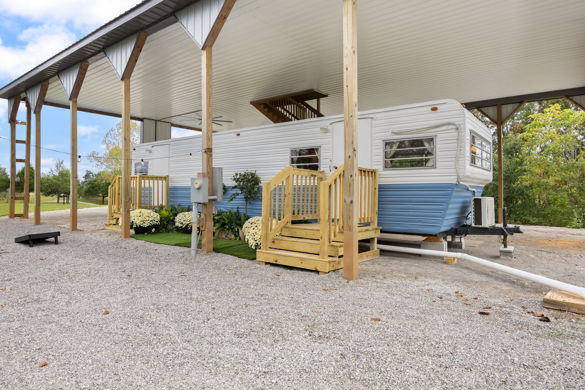 Vintage camper trailer with white and blue exterior parked under a large wooden carport with gravel ground and wooden steps leading to the doors.