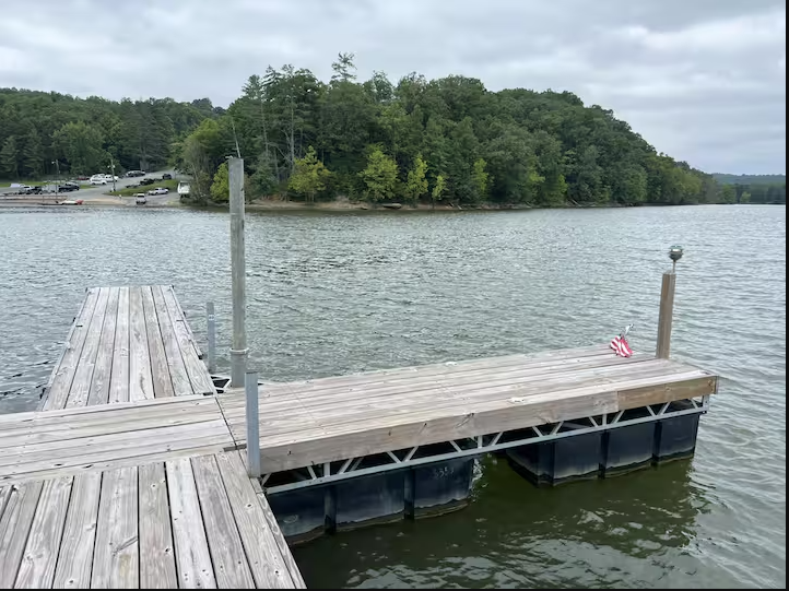 Wooden boat dock extending over calm lake water with forested hills reflected and a blue sky with scattered clouds.