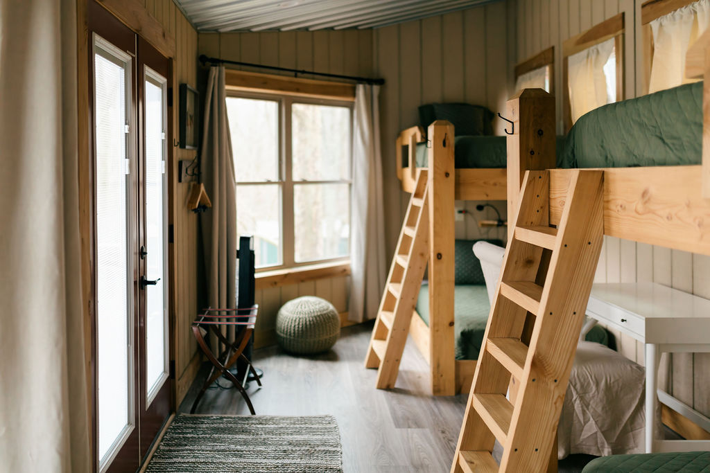 Bright A-frame loft room with wooden floors, a blue-covered sofa, large triangular window with blue curtains, and ceiling fan.