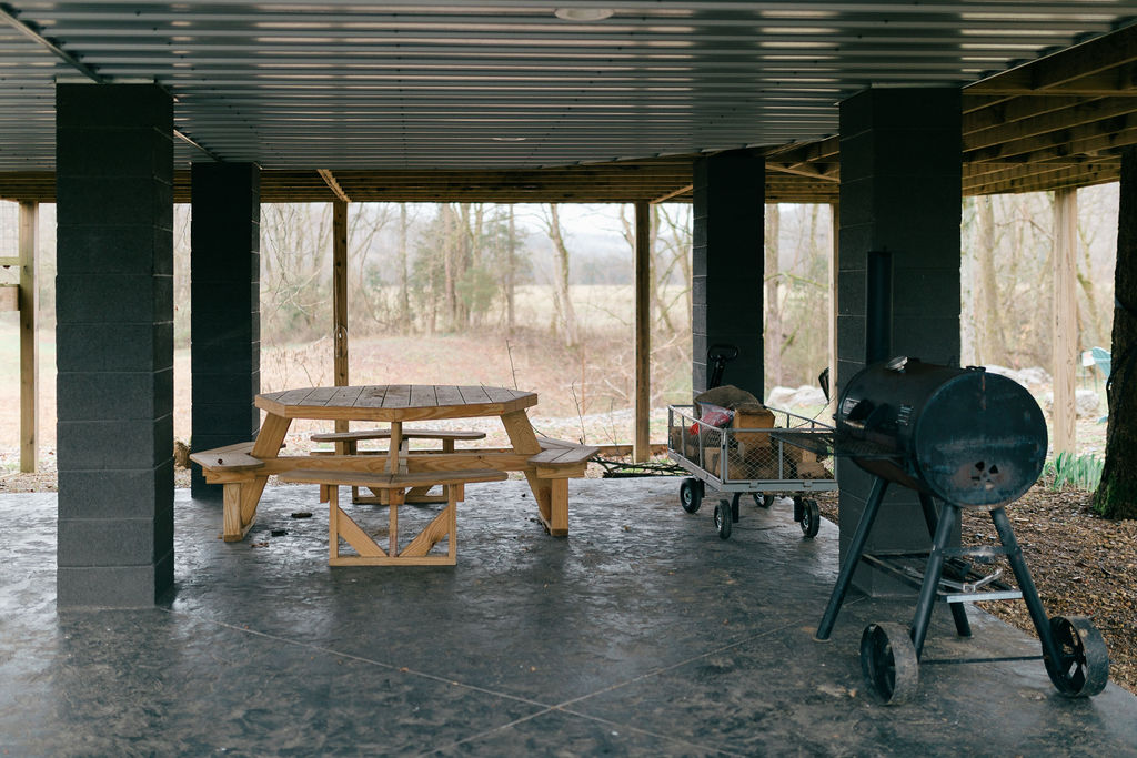 Modern bedroom with black wood bed frame, white bedding, green and sun patterned pillows, rug, window with gray curtains, standing fan, and wooden console with baskets and a potted plant.