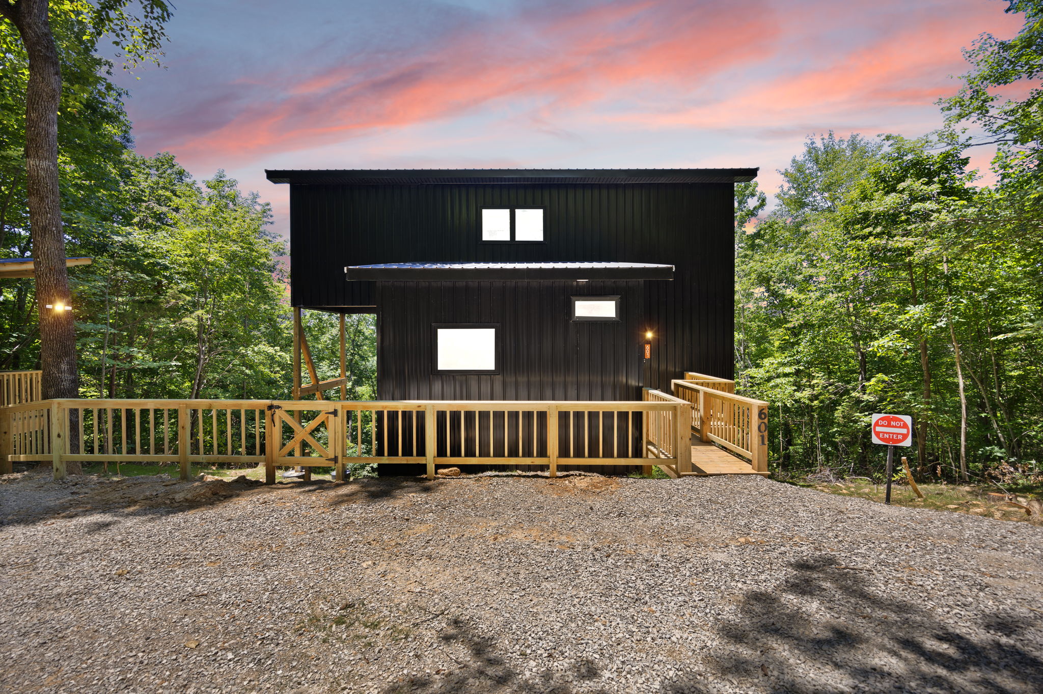 Modern black A-frame cabin with large glass windows and surrounding deck illuminated by small landscape lights at sunset.