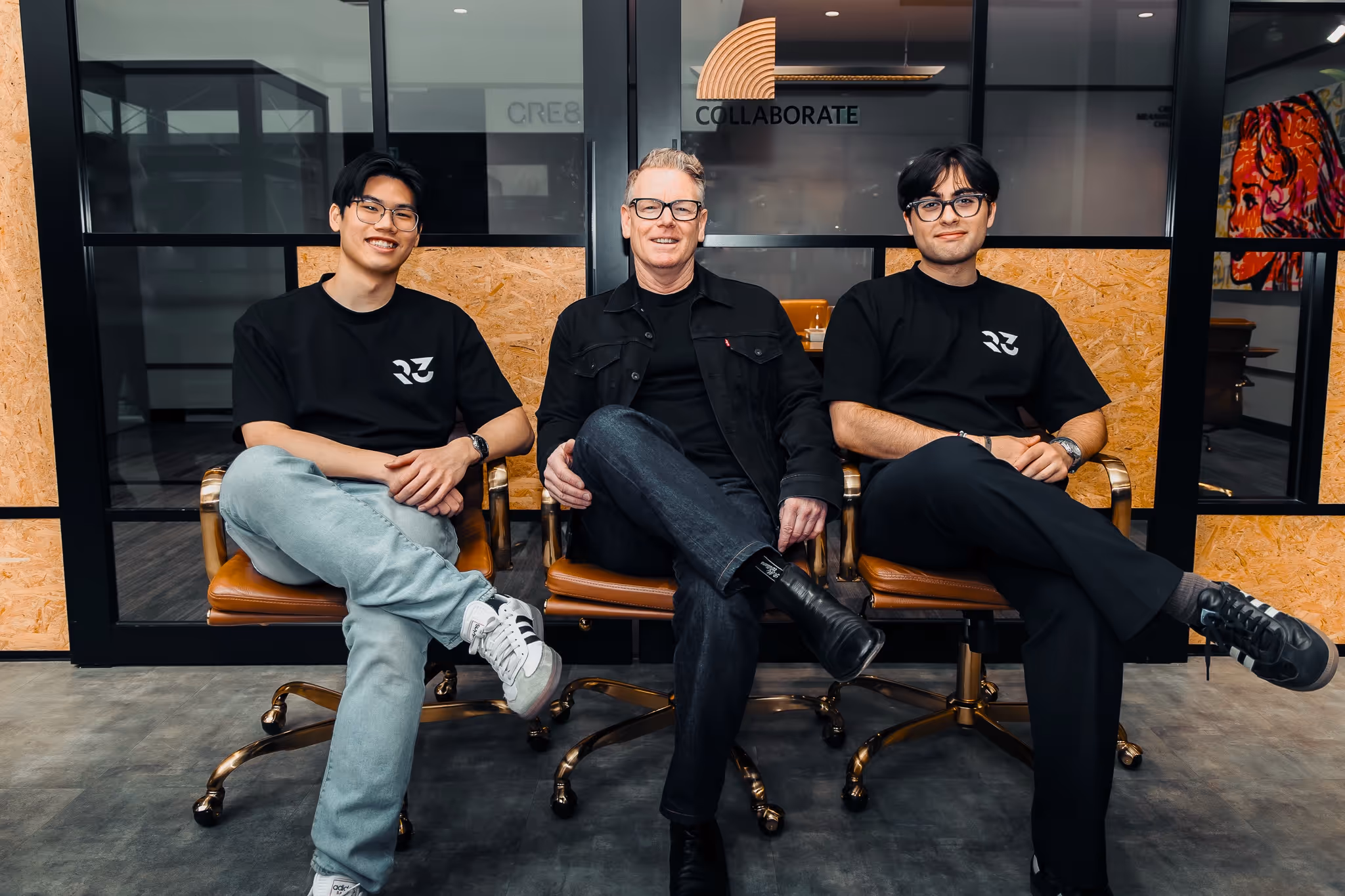 Three men sitting cross-legged on brown office chairs in front of a glass wall with the word 'COLLABORATE' and a logo above them.