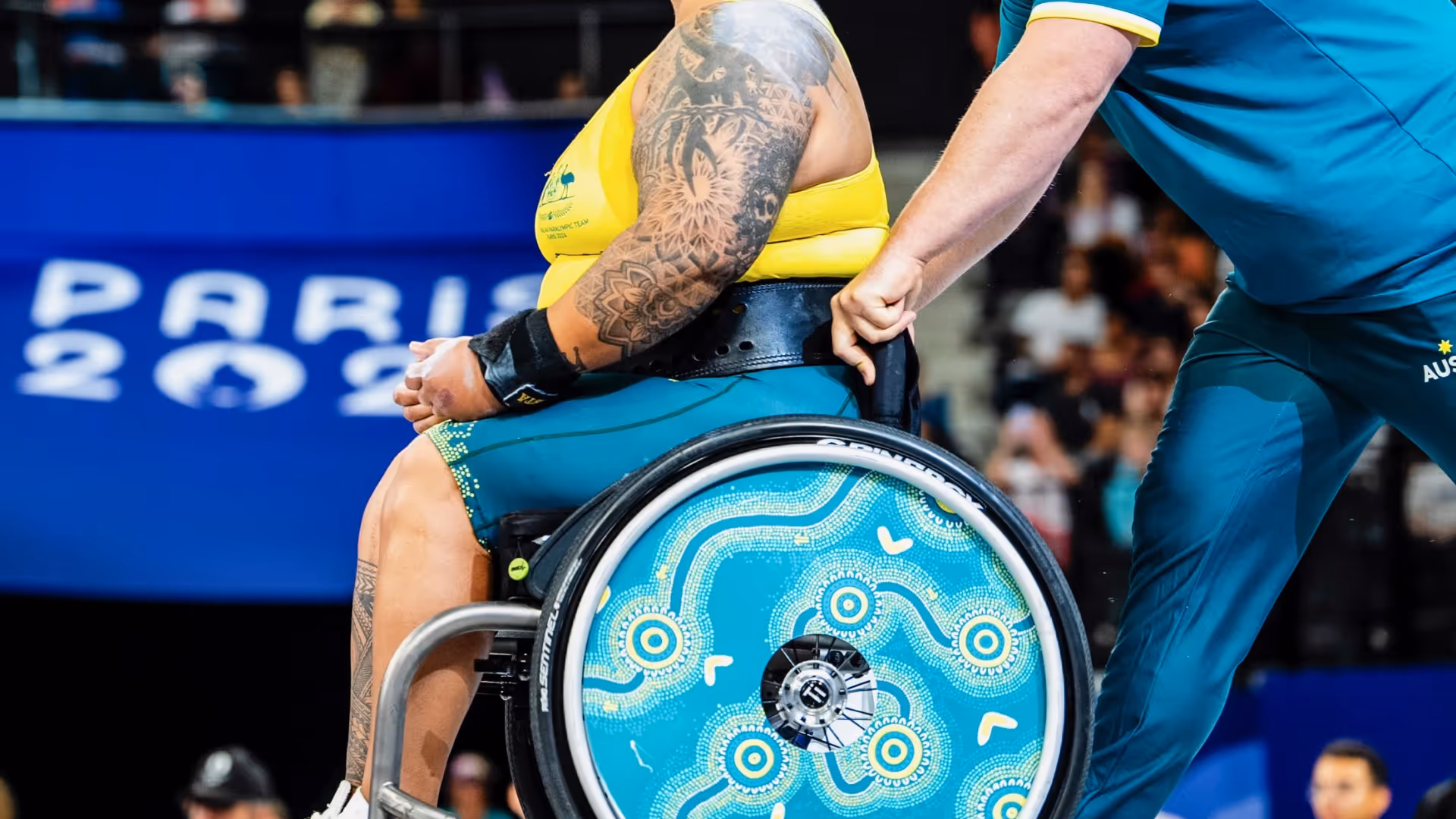 Athlete in a wheelchair with blue and yellow design being pushed by a person in teal sportswear at a Paris 2024 event.