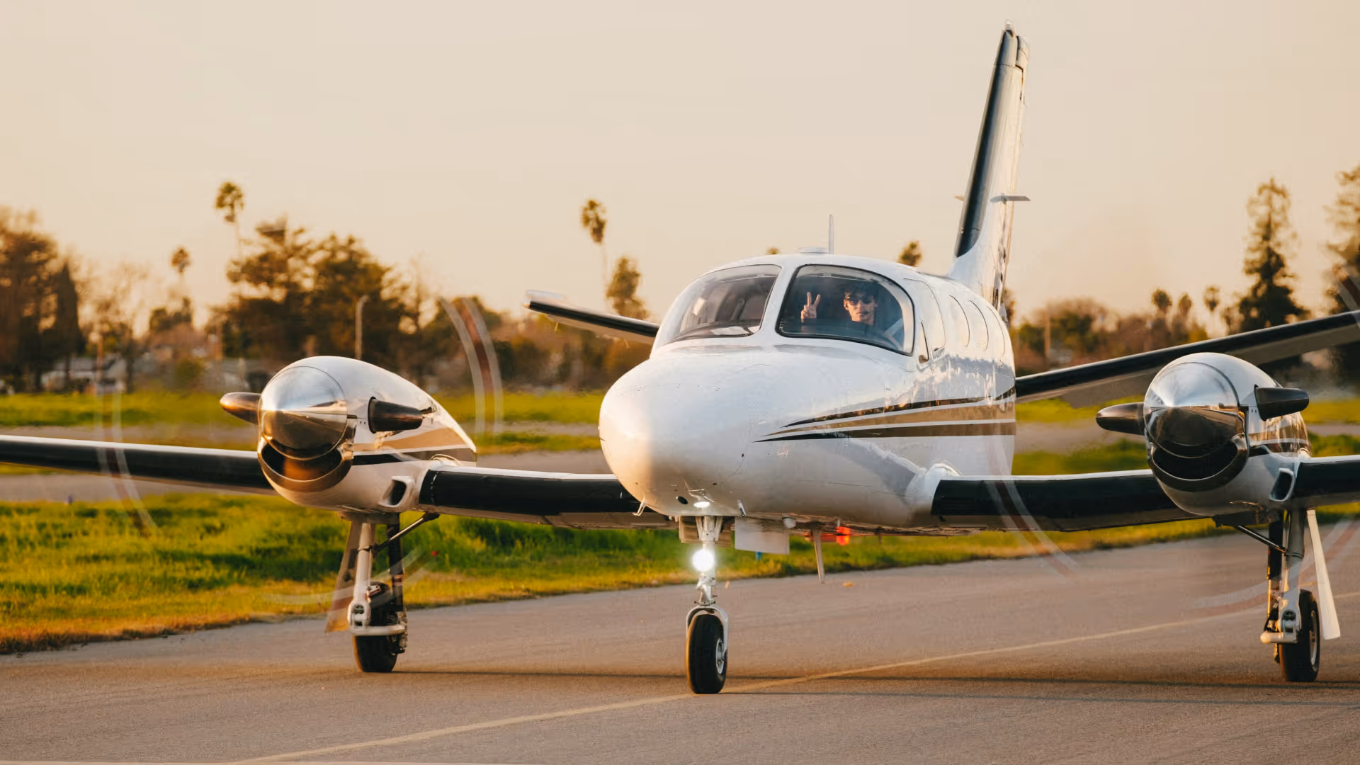 Small white twin-engine airplane taxiing on runway with pilot showing peace sign through window.