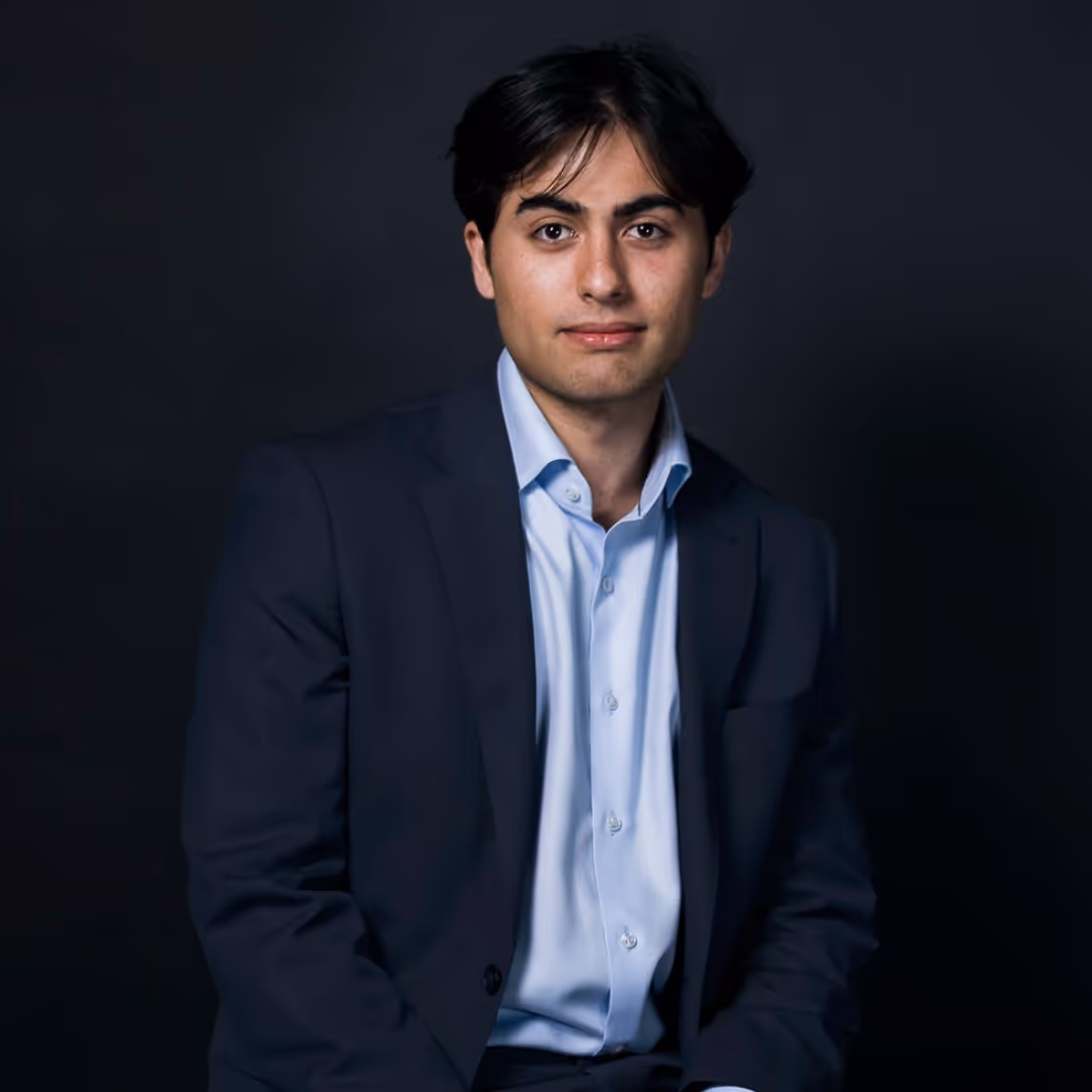 Young man with dark hair wearing a navy blazer and light blue shirt against a dark background.