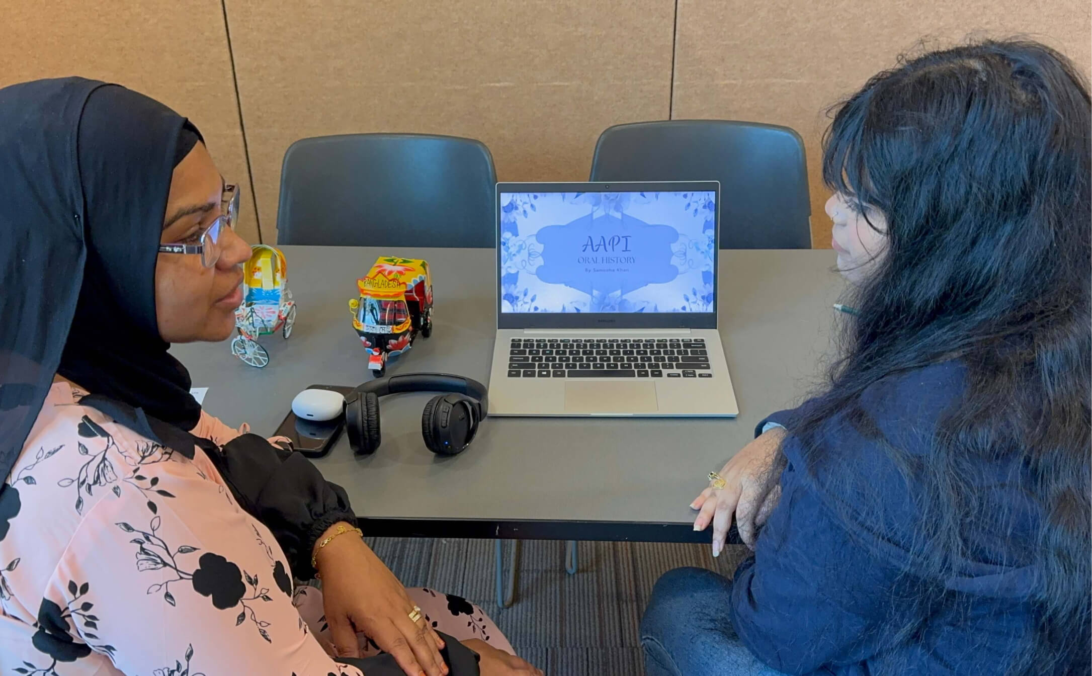 Two women sitting at a table looking at a laptop screen displaying a presentation titled 'AAPI Oral History.'
